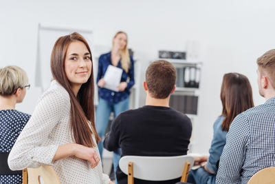 Bild vergr��ern: Young woman in a lecture or business presentation turning to smile at the camera viewed from the back of the room